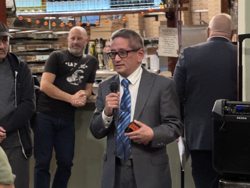 A man in a suit and tie speaks into a microphone at an indoor event, with people standing and observing in the background.