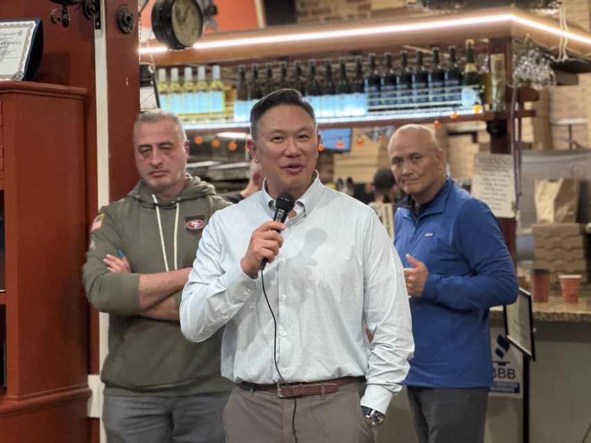 A man speaks into a microphone while two men stand behind him in a casual indoor setting with shelves and bottles in the background.