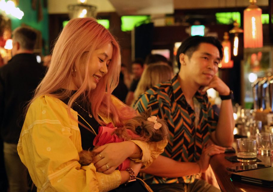 A woman with pink hair holds a small dog dressed in pink, while a man sits next to her at a bar with drinks and people in the background.