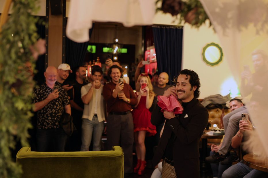 A man stands smiling and holding a pink cloth while a group of people in the background watch and take photos inside a warmly lit room.