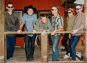 Six men wearing casual shirts and sunglasses stand and lean on a wooden porch railing in front of a red building, smiling and posing for the photo.