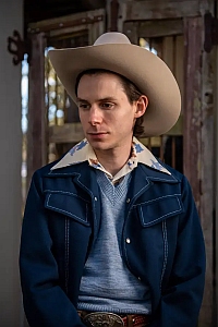 A young man in a cowboy hat and blue western jacket stands indoors, looking slightly downward with a neutral expression.