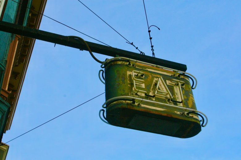 A weathered metal sign with the word "EAT" hangs from a pole against a blue sky.