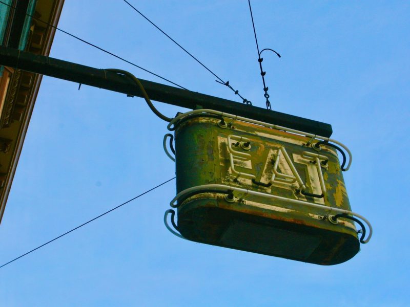 A weathered metal sign with the word "EAT" hangs from a pole against a blue sky.