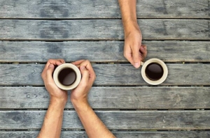 Two people holding cups of coffee sit across from each other at a wooden table viewed from above.