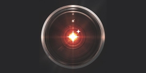 Close-up of a camera lens with a bright red light in the center, resembling a glowing eye, against a dark background.