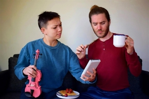 Two people sit on a couch; one holds a pink violin and a plate of food, the other holds a mug and notebook. Both appear to be reading the notebook with puzzled expressions.