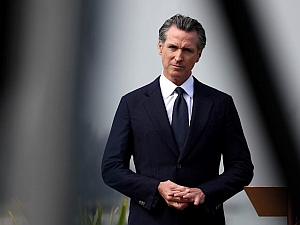 A man in a dark suit and tie stands outdoors near a podium, looking forward with his hands clasped.