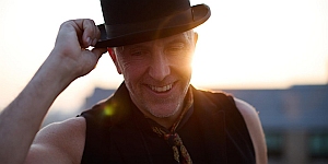 A person wearing a black top hat and vest smiles outdoors in the sunlight, holding the brim of the hat with one hand.