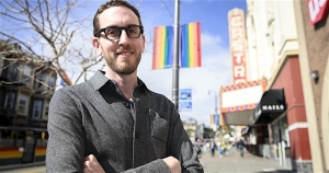 A man with glasses stands smiling on a city street near a rainbow pride flag and the Castro Theatre in the background.