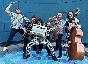 Five men pose with musical instruments, including a drumstick, keyboard, violin, and double bass, standing on a blue patterned floor against a blue tiled wall.