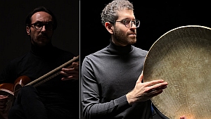 Two men in dark clothing: one plays a string instrument in low light, the other holds a large round percussion instrument against a dark background.