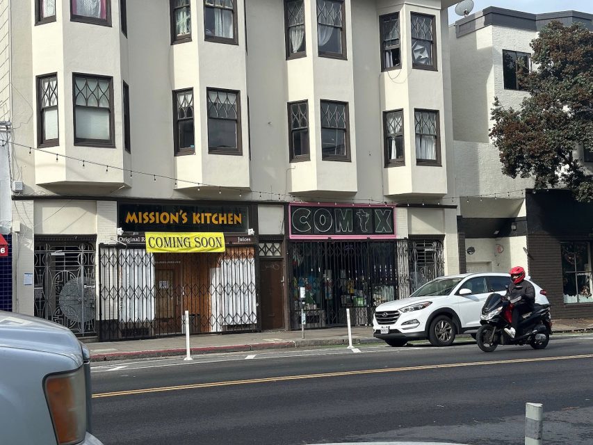 Street view of a building with signs for “Mission’s Kitchen Coming Soon” and “Comix”; a person on a scooter and a white SUV are on the road in front.