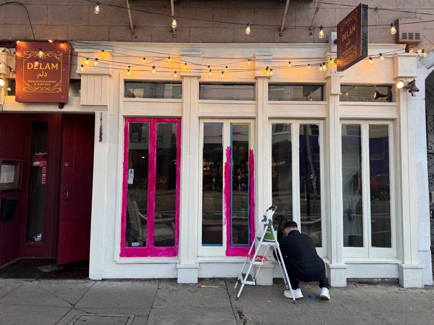 A person paints the lower panels of a storefront window bright pink while sitting on a small ladder outside a shop called DéLAM.