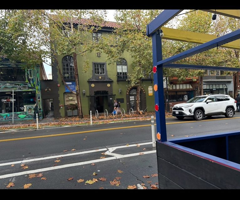 A white SUV is parked on a city street lined with trees and buildings; a person stands on the sidewalk, and autumn leaves cover the ground.