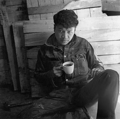 A person in a denim jacket sits on the ground against wooden planks, holding a cup and looking toward the camera.