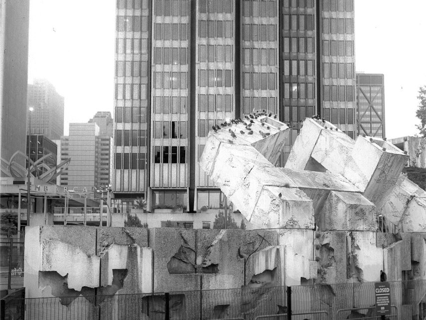 A large, abstract concrete sculpture in disrepair stands in front of tall office buildings in an urban setting, with a "closed" sign visible on a barrier.