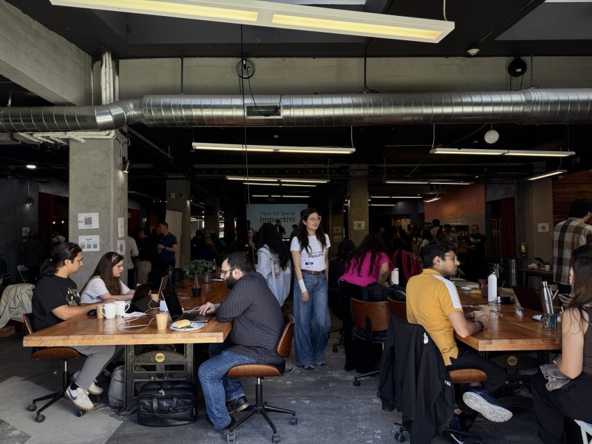 Open-plan office space with people working at shared wooden tables on laptops; some are talking, and a few are standing or walking through the room.