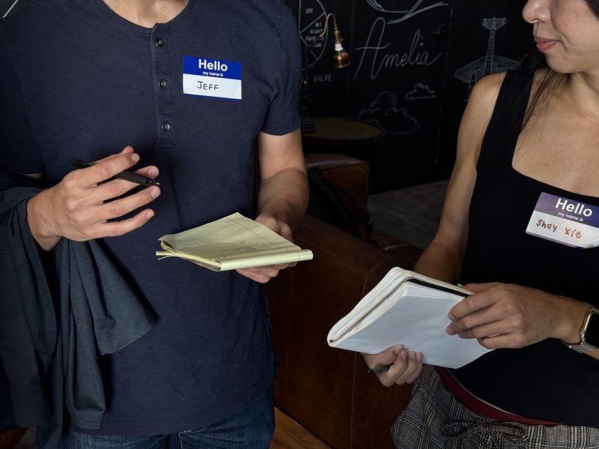 Two people wearing name tags, "Jeff" and "Shay Xie," stand indoors holding notebooks and pens, appearing to have a conversation.