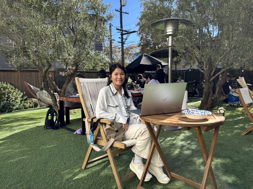 A person in light clothing sits on a wooden chair outdoors with an open laptop on a round table, surrounded by greenery and other people at nearby tables.