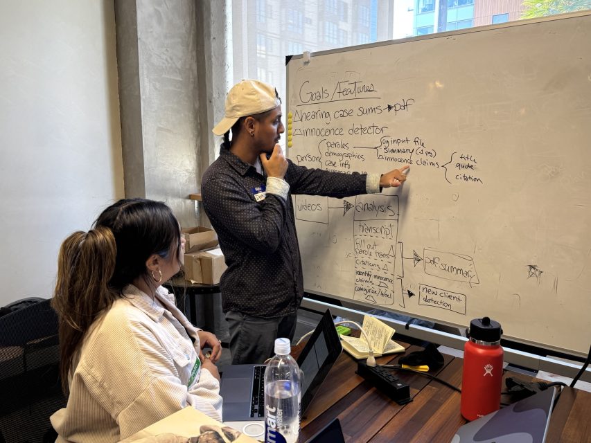 Two people collaborate in an office, with one person pointing at notes and diagrams on a whiteboard about goals, thresholds, and case analysis, while the other observes.
