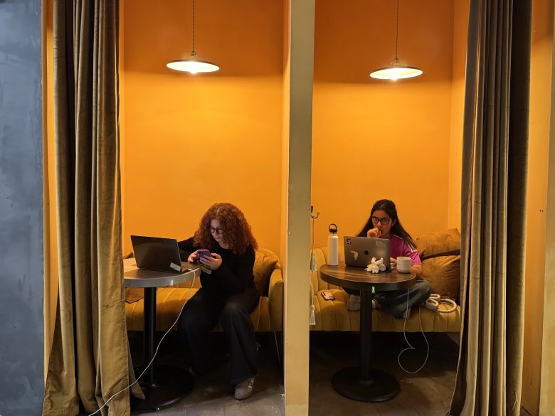 Two women work on laptops in adjacent, curtained yellow booths; one checks her phone while the other looks at her screen, both with drinks on their tables.