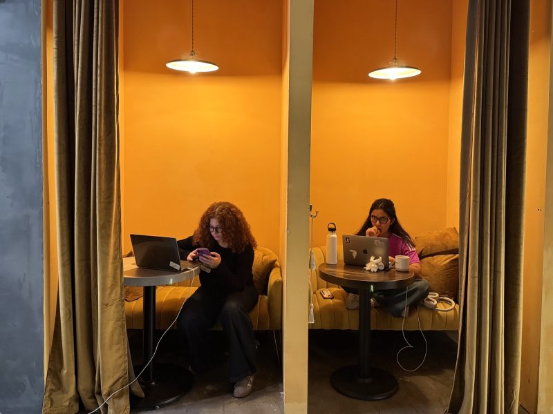 Two women work on laptops in adjacent, curtained yellow booths; one checks her phone while the other looks at her screen, both with drinks on their tables.