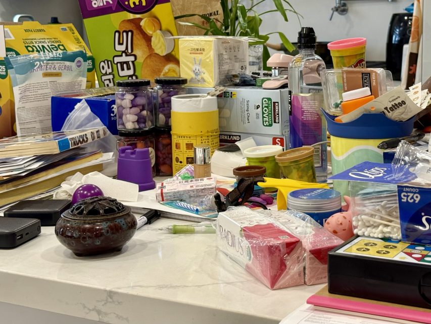 A cluttered countertop covered with snacks, containers, toys, stationery, and various household items in a kitchen or living space.