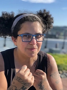 A person with curly hair in pigtails, wearing glasses and a headband, holds up their fists outdoors near water on a sunny day.