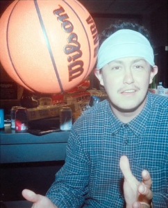 A man wearing a white headband and plaid shirt sits indoors with a large basketball labeled "Wilson NBA" floating near his head.