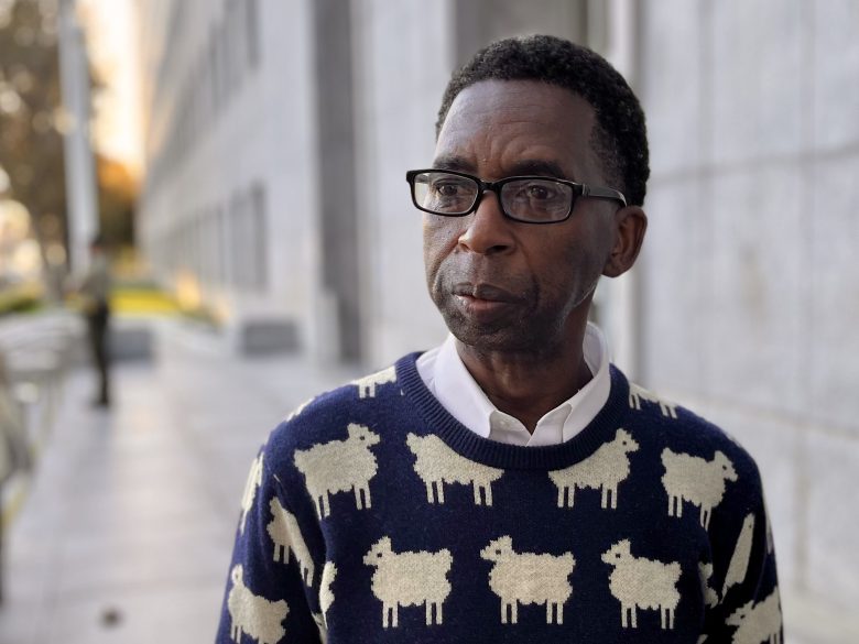 A man wearing glasses and a navy sweater with white sheep patterns stands outside near a modern building.