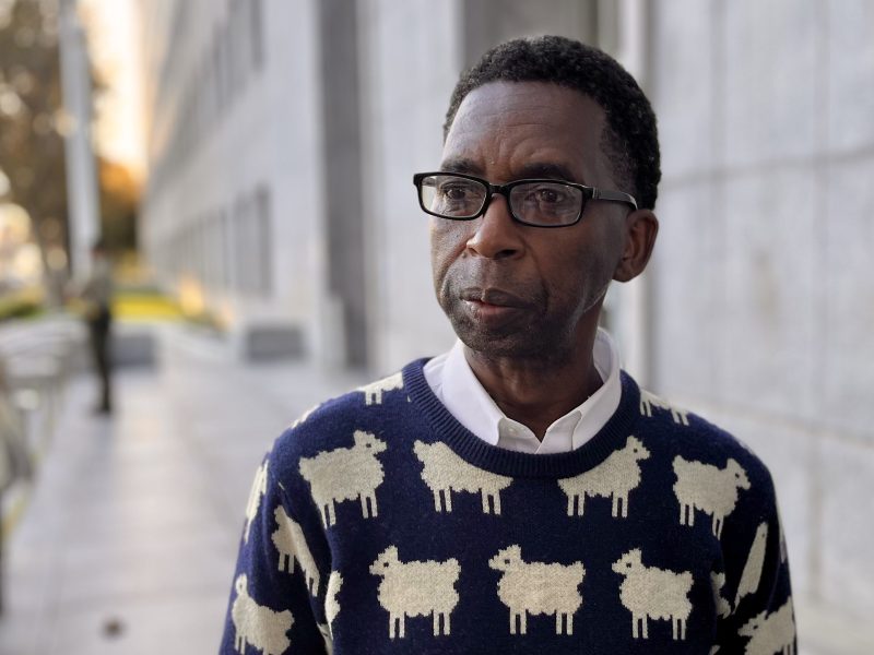 A man wearing glasses and a navy sweater with white sheep patterns stands outside near a modern building.