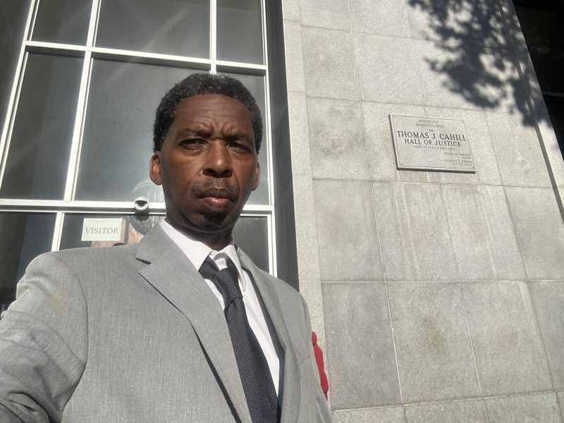 A man in a gray suit and tie stands outside a stone building near a window and a plaque reading "Thomas J. Cahill Hall of Justice.