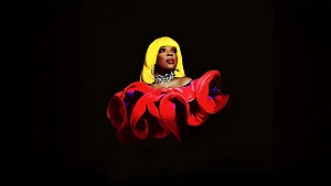 A person with a bright yellow wig and dramatic makeup wears a large, sculptural red ruffled garment against a black background.