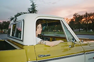 A person with long brown hair sits in the driver's seat of a vintage yellow and white vehicle, looking toward the camera at sunset.