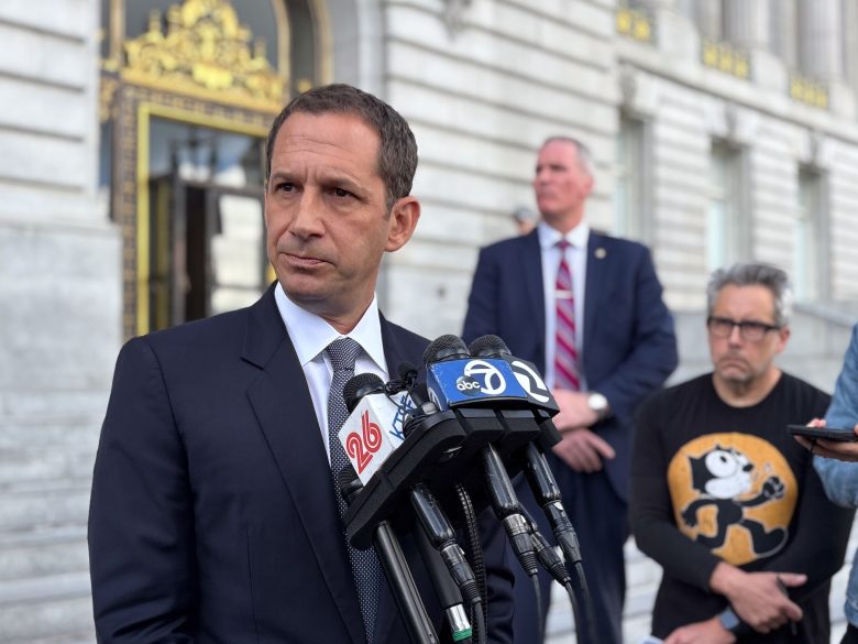 A man in a suit speaks at a podium with multiple microphones outside a building, while two other men stand in the background.