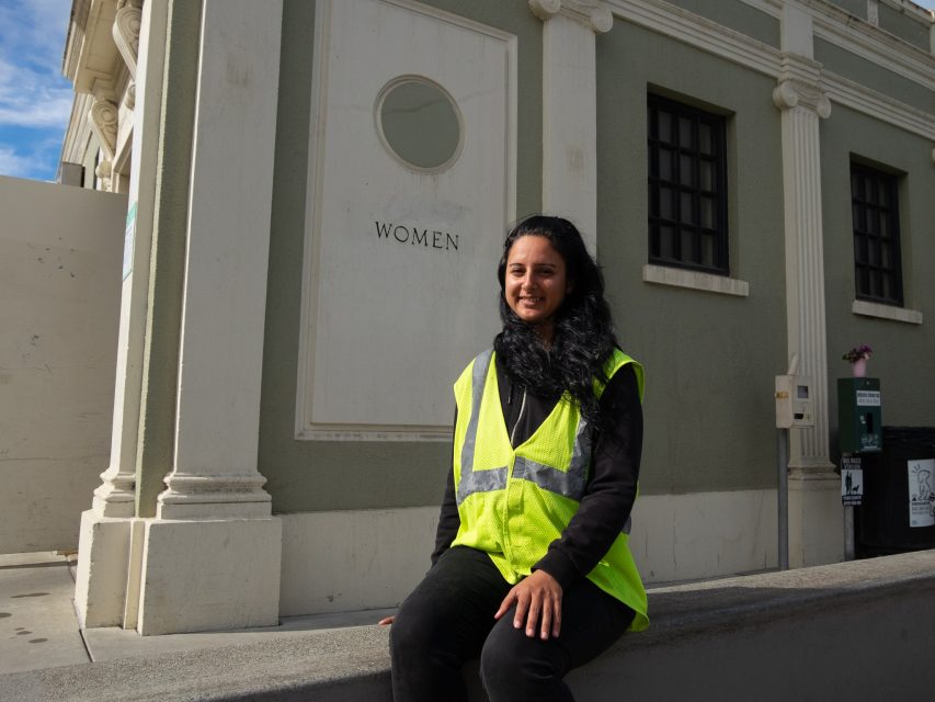 A person wearing a yellow safety vest sits on a low concrete wall in front of a building marked "WOMEN.