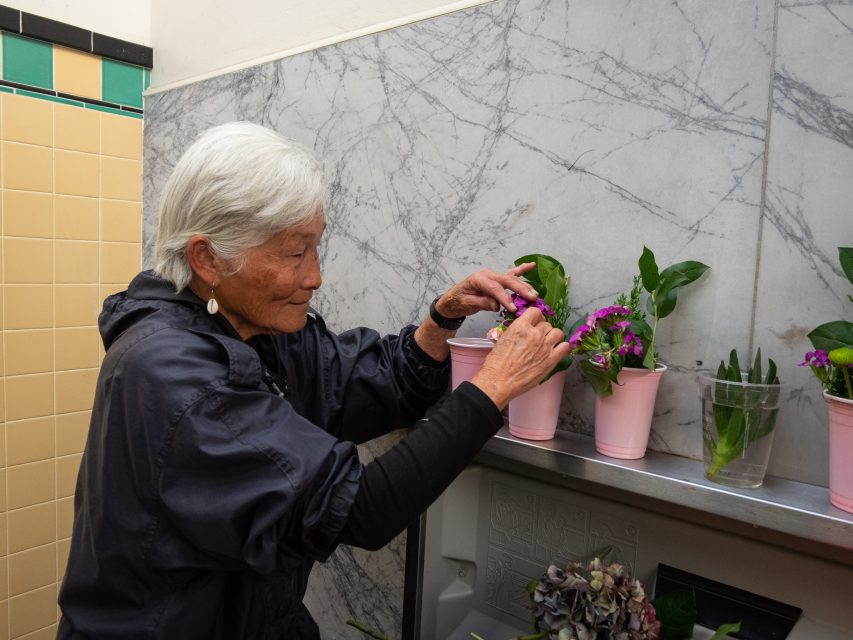 An older woman arranges purple flowers in pink pots on a shelf against a marble-patterned wall.