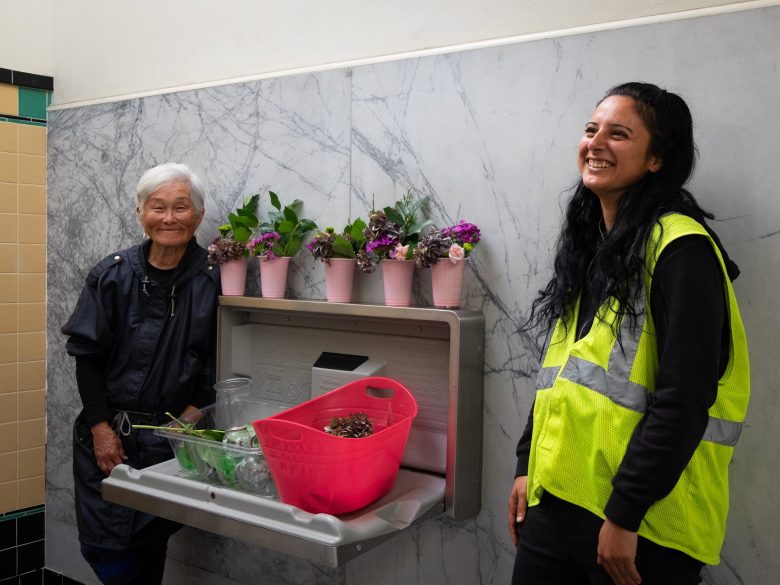 Two women stand by a wall-mounted changing table holding potted flowers and a pink bucket. One woman wears a safety vest and the other is in dark clothing. Both are smiling.