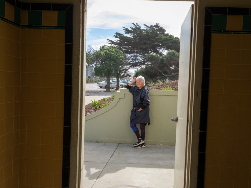 An older woman stands outside near a low wall, visible through an open doorway, with trees and parked cars in the background.