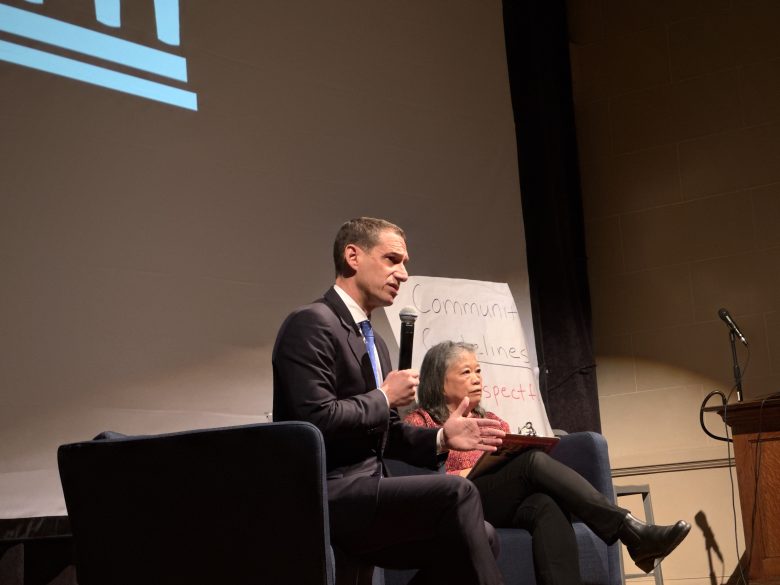 A man in a suit speaks into a microphone while seated next to a woman on stage; a whiteboard with handwritten words is in the background.