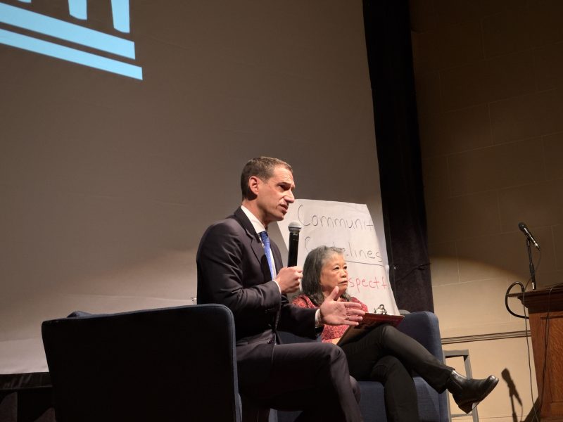 A man in a suit speaks into a microphone while seated next to a woman on stage; a whiteboard with handwritten words is in the background.