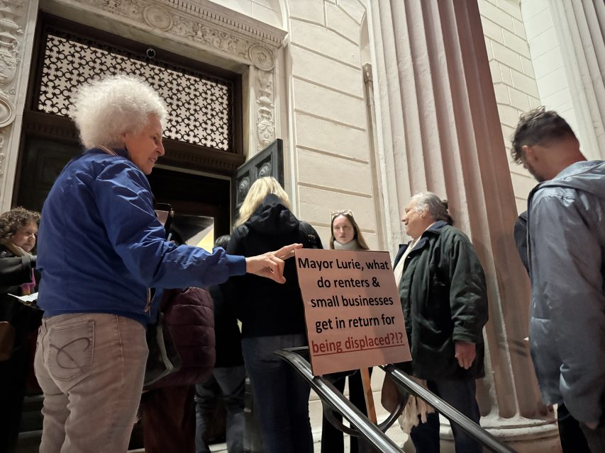 A person holds a sign asking the mayor what renters and small businesses get in return for being displaced while standing with others outside a building.
