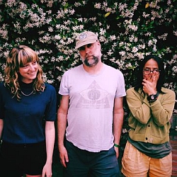 Three adults stand in front of a wall covered with white flowers; the person in the center wears a hat, while the others stand on either side, posing casually.