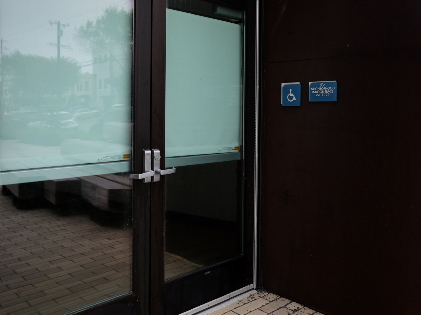 Glass door entrance with double handles, next to a blue wheelchair accessible sign on a dark wall; sidewalk and parked cars visible through the glass.