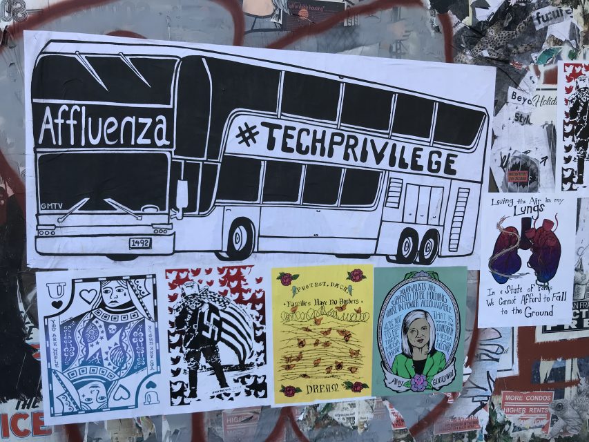 A black-and-white poster of a double-decker bus labeled "Affluenza" and "#TECHPRIVILEGE" stands out above colorful artwork and posters on an art wall covered in graffiti.