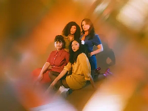 Four women pose together against a warm, softly blurred background, each wearing different colored outfits. The image is partially obscured by an abstract, hazy overlay.