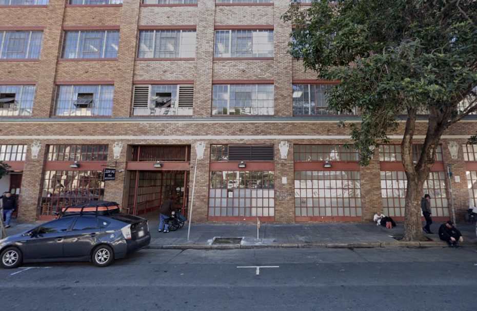 A brick building with large windows and garage doors lines a city street; several people are standing or sitting nearby, and a parked car is visible in the foreground.