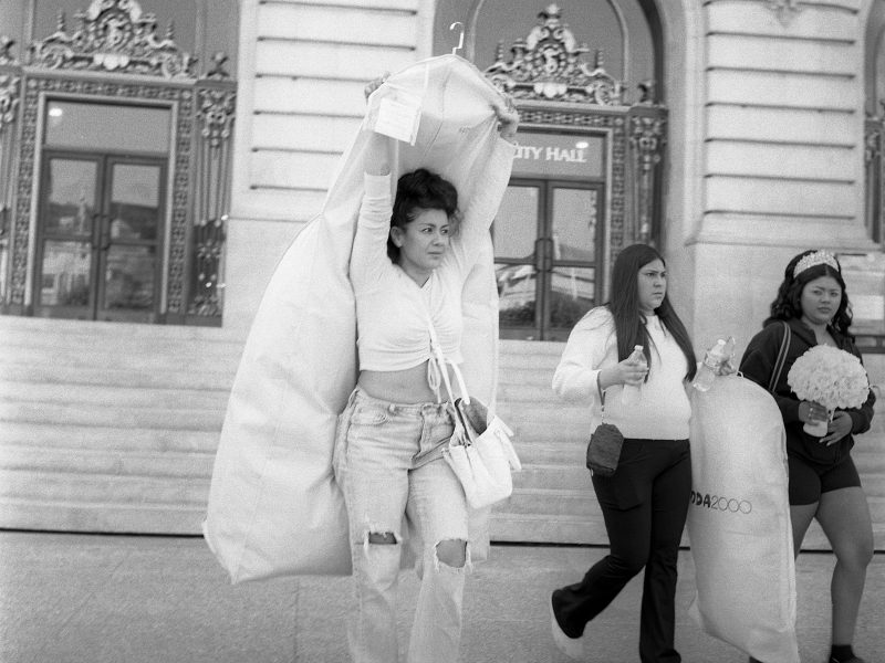 Three women walk outside a building; the woman in front carries a garment bag over her head, while the others hold items including what appear to be flowers and another garment bag.