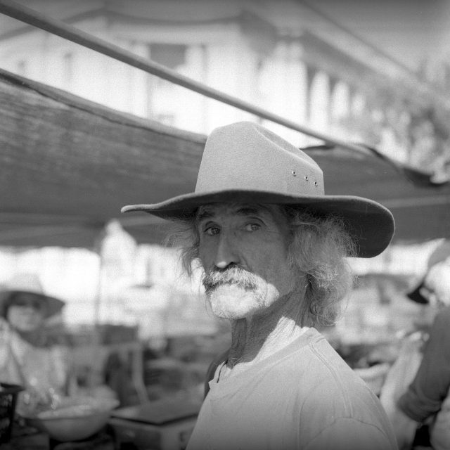An older man with a large brimmed hat stands outdoors at a market, looking towards the camera. The image is in black and white.